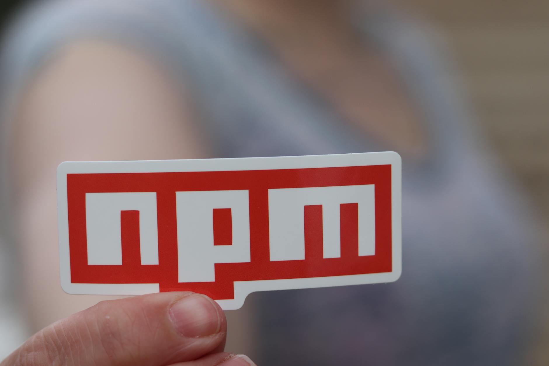 Dłoń trzymająca naklejkę npm na rozmytym tle developerskim